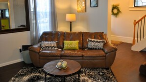 A living room with a brown leather sofa, patterned cushions, a round coffee table with decorative items, a floor lamp, and a staircase. - Oswego Downtown District (Oswego)