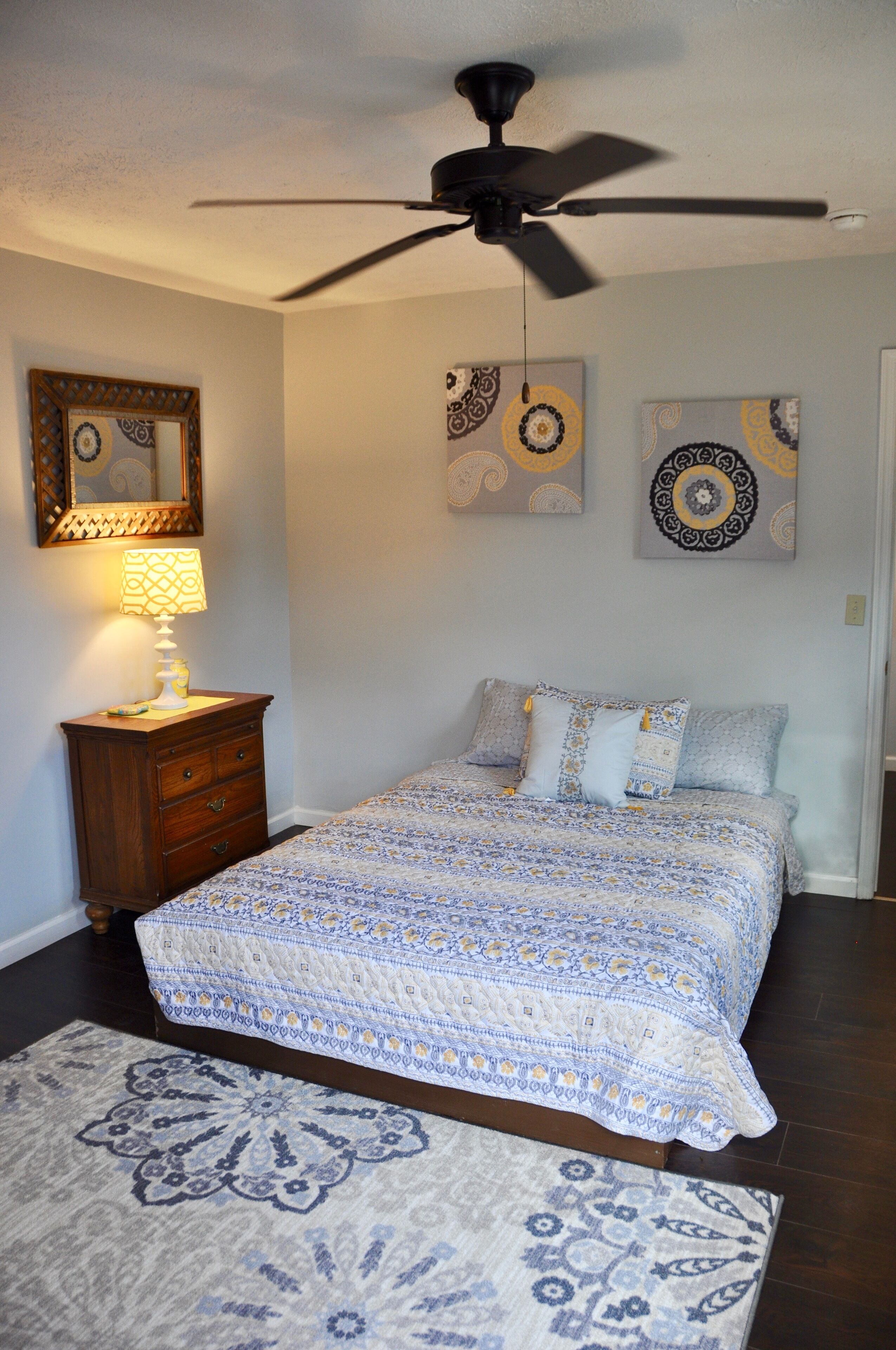 A bedroom with a bed, a ceiling fan, a dresser, a lamp, and wall art.