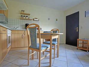 Een compacte keuken met houten kastjes, een inbouwoven en een betegelde spatwand. Er staan twee houten stoelen met blauwe kussens rond een kleine tafel.