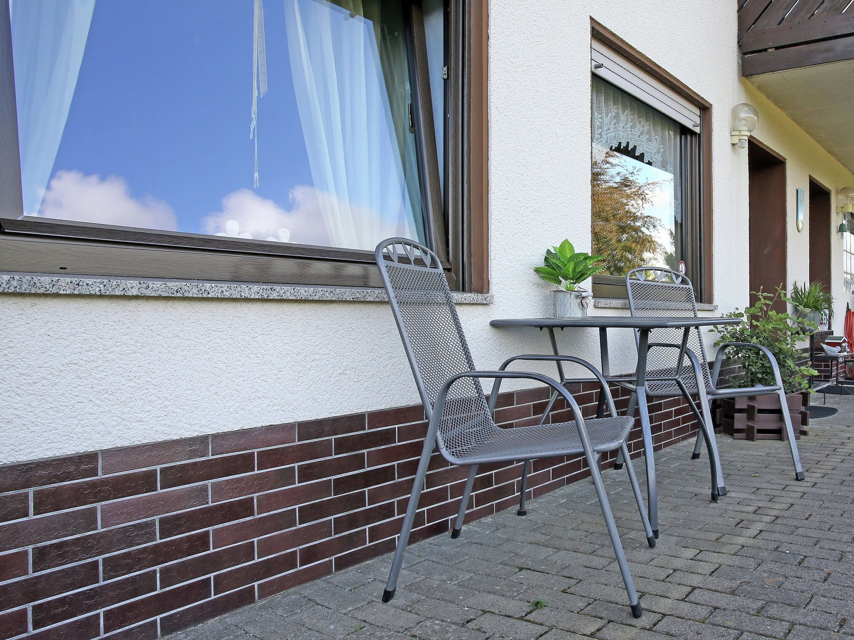 An outdoor seating area with metal chairs and a table, a brick wall, and a window with a curtain.