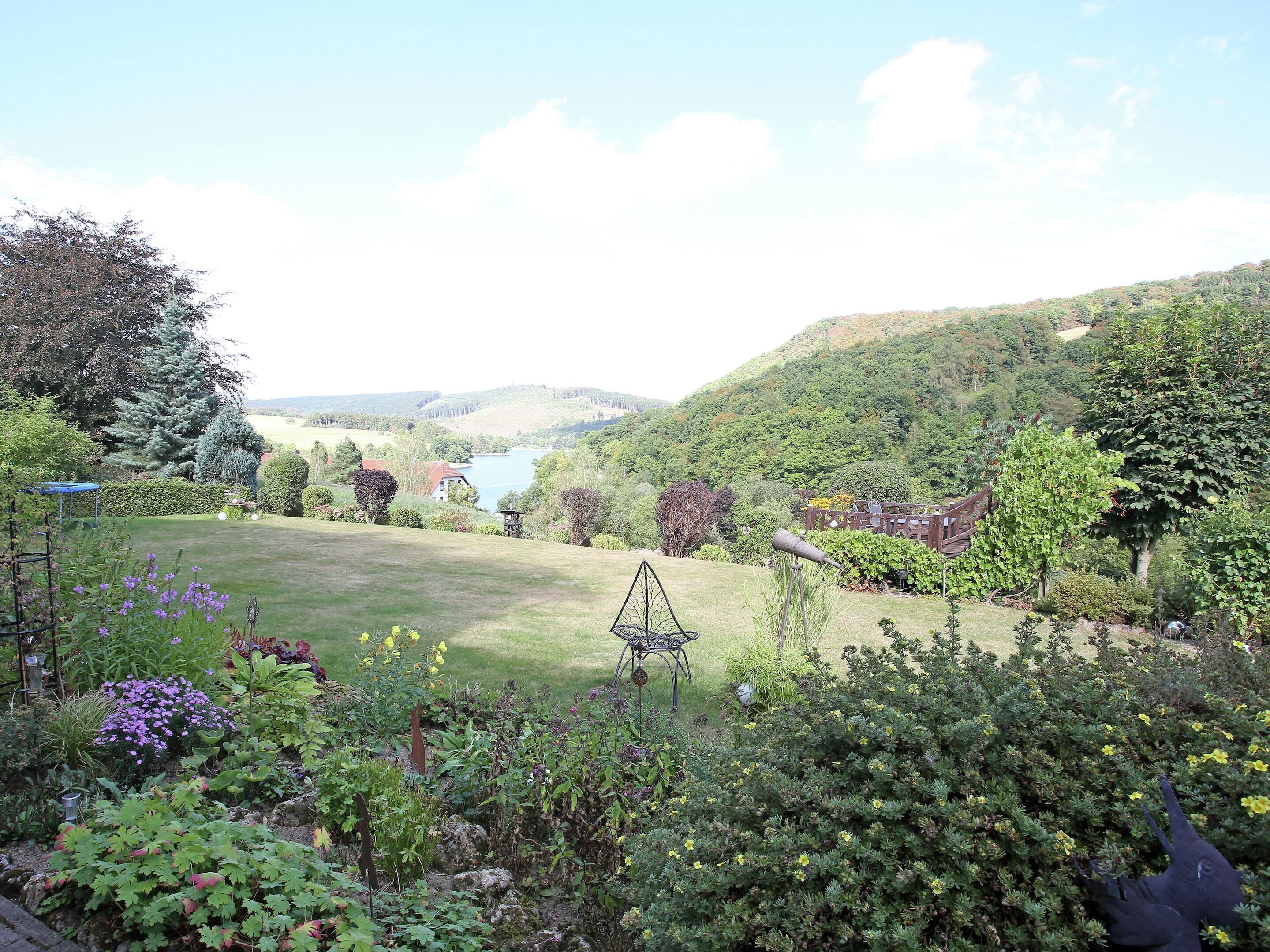 Un jardín con césped, diversas plantas, un telescopio y vistas a un lago y colinas al fondo.