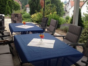 Ein Sitzbereich im Freien mit Tischen und Stühlen, umgeben von Grünflächen und Gebäuden im Hintergrund.