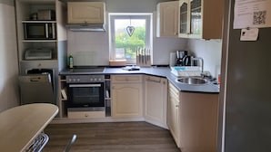 Eine kompakte Küche mit Mikrowelle, Backofen und Geschirrspüler. Ein Fenster mit Herzdeko, und ein QR-Code am Kühlschrank.