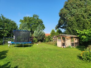 Un patio trasero con un trampolín, una pequeña estructura de madera y abundante vegetación.
