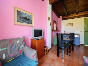 Un salon avec un mur rose, un téléviseur posé sur un meuble en bois, un canapé agrémenté de coussins colorés, une table à manger avec des chaises et une porte verte.