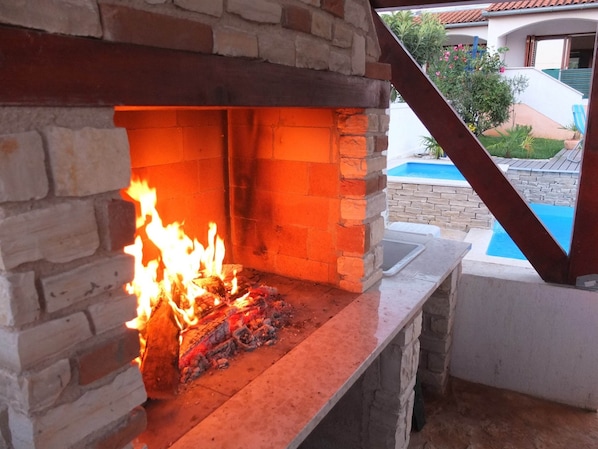 A stone fireplace with a fire burning inside, a pool, and a view of a house in the background.