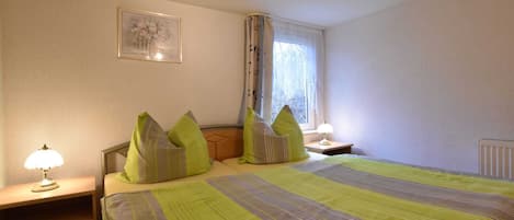 A bedroom with a bed, two bedside tables, a lamp, a window with curtains, and a framed picture on the wall.