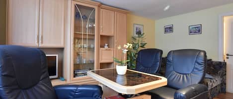 Una sala de estar con una mesa de comedor, dos sillones de cuero azul, un aparador con puertas de cristal y una planta en maceta.