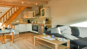 Uma sala de estar acolhedora com piso de madeira, um sofá cinza, uma mesa de centro de madeira e uma escada que leva a uma cozinha com armários brancos e um fogão.
