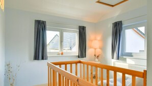 Quarto com corrimão de madeira, duas janelas com cortinas quadriculadas em preto e branco, luminária de cabeceira e radiador.