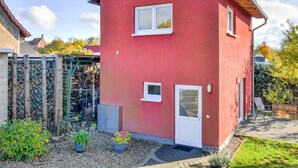 Uma casa vermelha de dois andares com um pequeno alpendre, porta de garagem visível e um jardim com plantas em vasos.