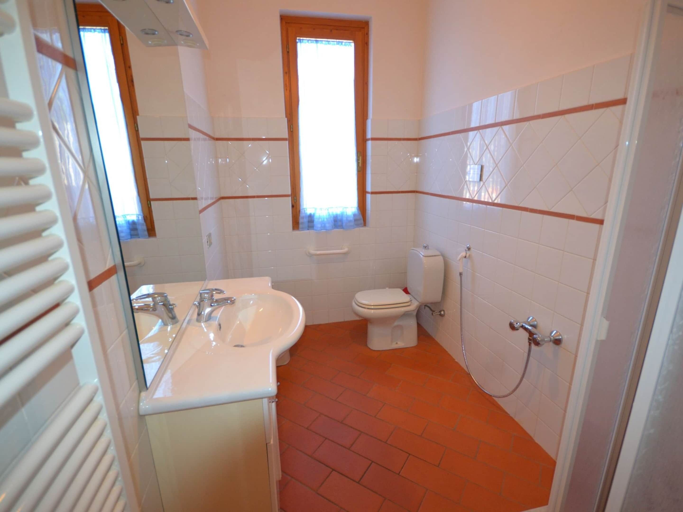 Un baño con un lavamanos blanco, espejo e inodoro. Hay una ventana sobre el inodoro y un radiador a la izquierda.