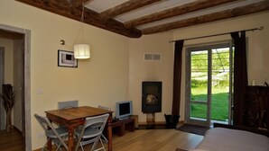 Ein Zimmer mit Holzbalkendecke, einem Esstisch mit Stühlen, einem Fernseher, einem Kamin und einer großen Schiebeglasdoor zu einem Garten.