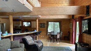 A cozy cabin interior with a sofa, a rug, a dining table with chairs, a kitchen area, and a TV.