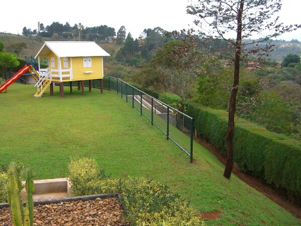 A yellow playhouse with a slide and stairs, surrounded by a fenced area with a grassy lawn and a cactus in the foreground.