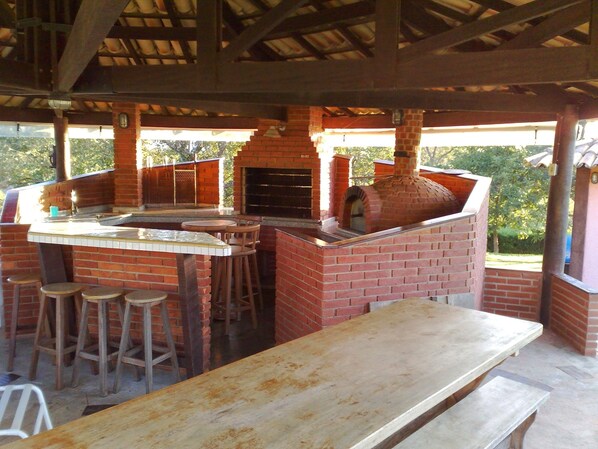 Een buitenkeuken met een bakoven, barstoelen en een lange tafel.