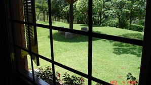 A view through a window with metal bars, featuring a grassy area with a thatched structure, trees, and a stone bench. - Chácara Vale da Esperança 40 minutes from São Paulo with Fiber Internet (Mairinque)
