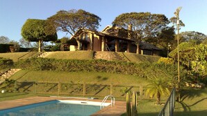 A pool area with a clear view of a house with a stone facade and a sloped roof, surrounded by a well-maintained garden and a fence. - Chácara Vale da Esperança 40 minutes from São Paulo with Fiber Internet (Mairinque)