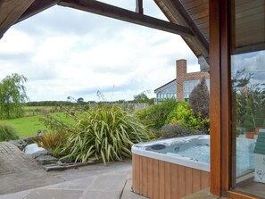 Un bain à remous dans un jardin agrémenté d’une pergola en bois, une maison en briques en arrière-plan et un champ verdoyant.