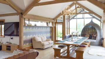 A spacious room with a high ceiling, wooden beams, a chandelier, a large window, a sofa, a dining table, and a seating area with cushions.
