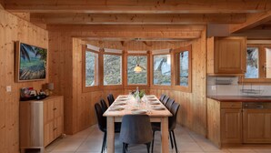 A wooden-paneled dining room with a large table set for eight, flanked by chairs, and a kitchen area with wooden cabinets and a window. - Chalet Belle Roche | Recently renovated | Jacuzzi (Riddes)