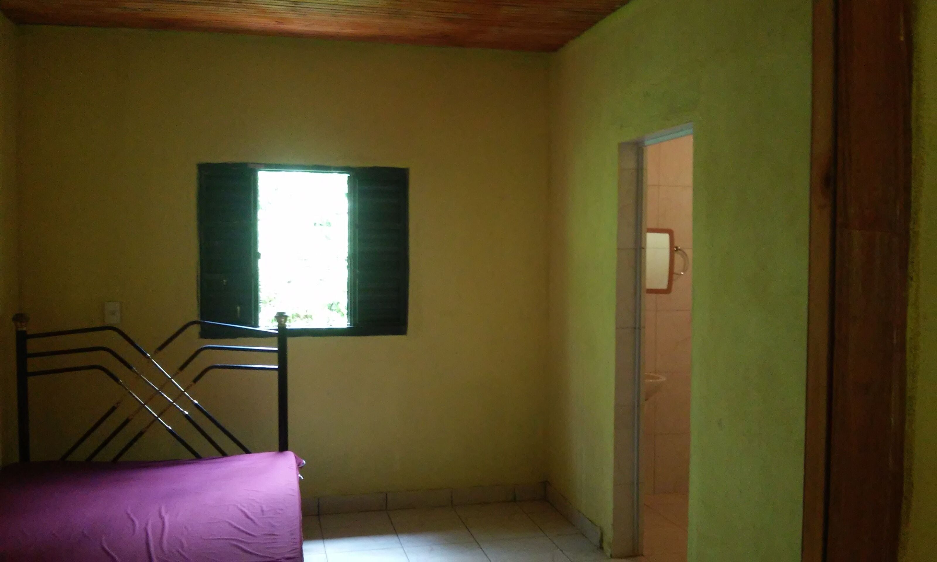 A small, dimly lit room with a single window, a metal bed frame, and a door leading to another room.