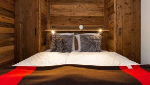 A wooden room with a bed, two fluffy pillows, a clock, and a red and black blanket.