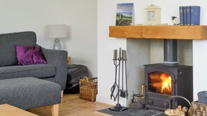 A cozy living room with a grey sofa, a purple cushion, a lamp, a bookshelf with books, a fireplace, and a basket of firewood. - 4 bedroom accommodation in Goathland (Goathland)
