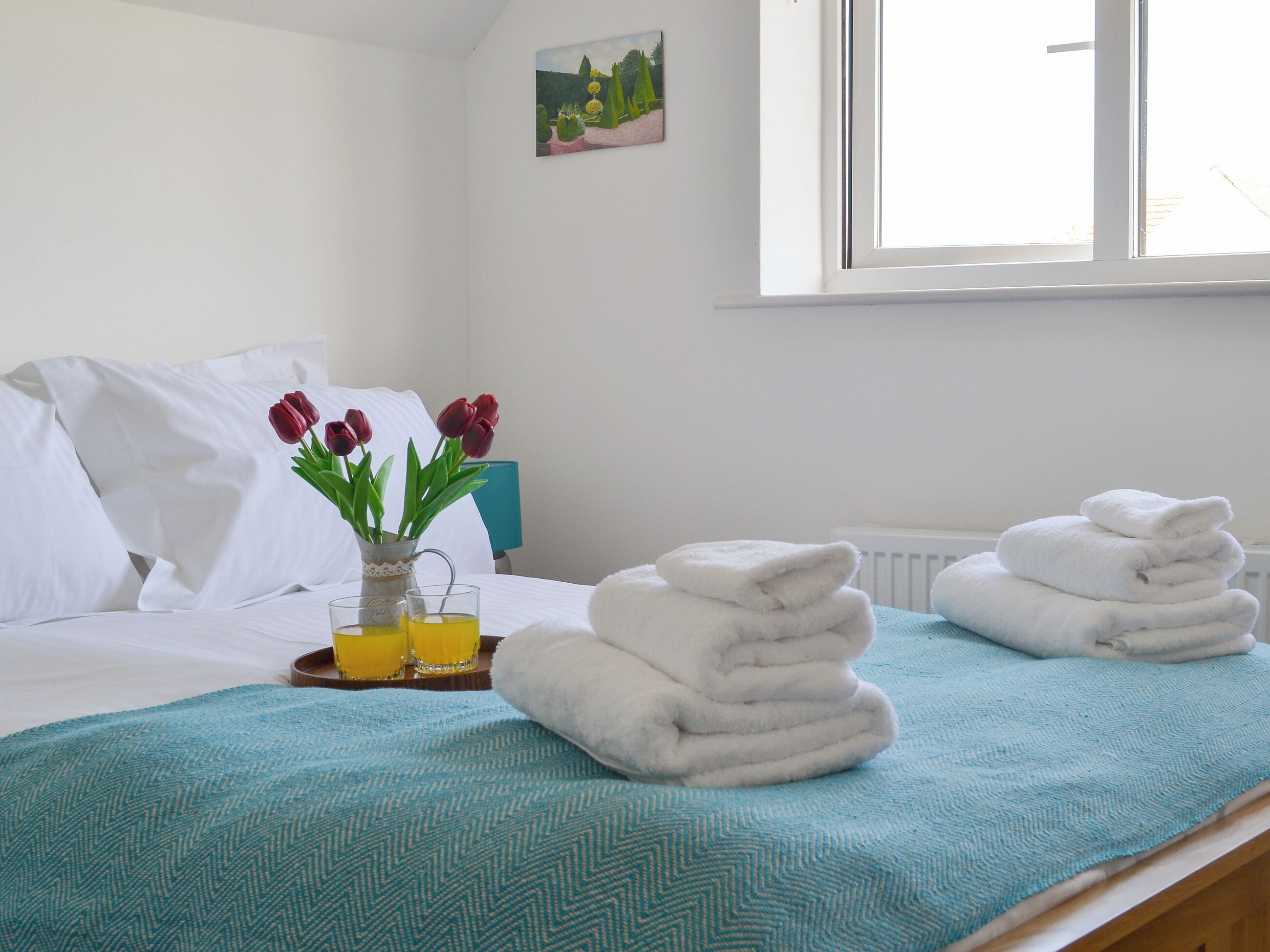 A neatly made bed with white linens, a teal bedspread, and a stack of white towels. There is a vase with red tulips and a glass of yellow liquid on a tray beside the bed.