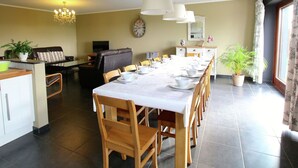 Ein Essbereich mit einem langen Tisch, der für eine Mahlzeit gedeckt ist, umgeben von Holzsesseln. Im Hintergrund befindet sich ein Fernseher, eine Chandelüster hängt von der Decke und eine Uhr ist an der Wand angebracht.