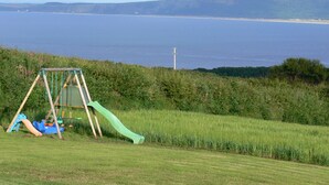 A grassy area with a children's playset including a slide and swings, overlooking a large body of water and distant hills. - RUSTIC LOG CABIN LOCATED GREENCASTLE CO DONEGAL (Greencastle)