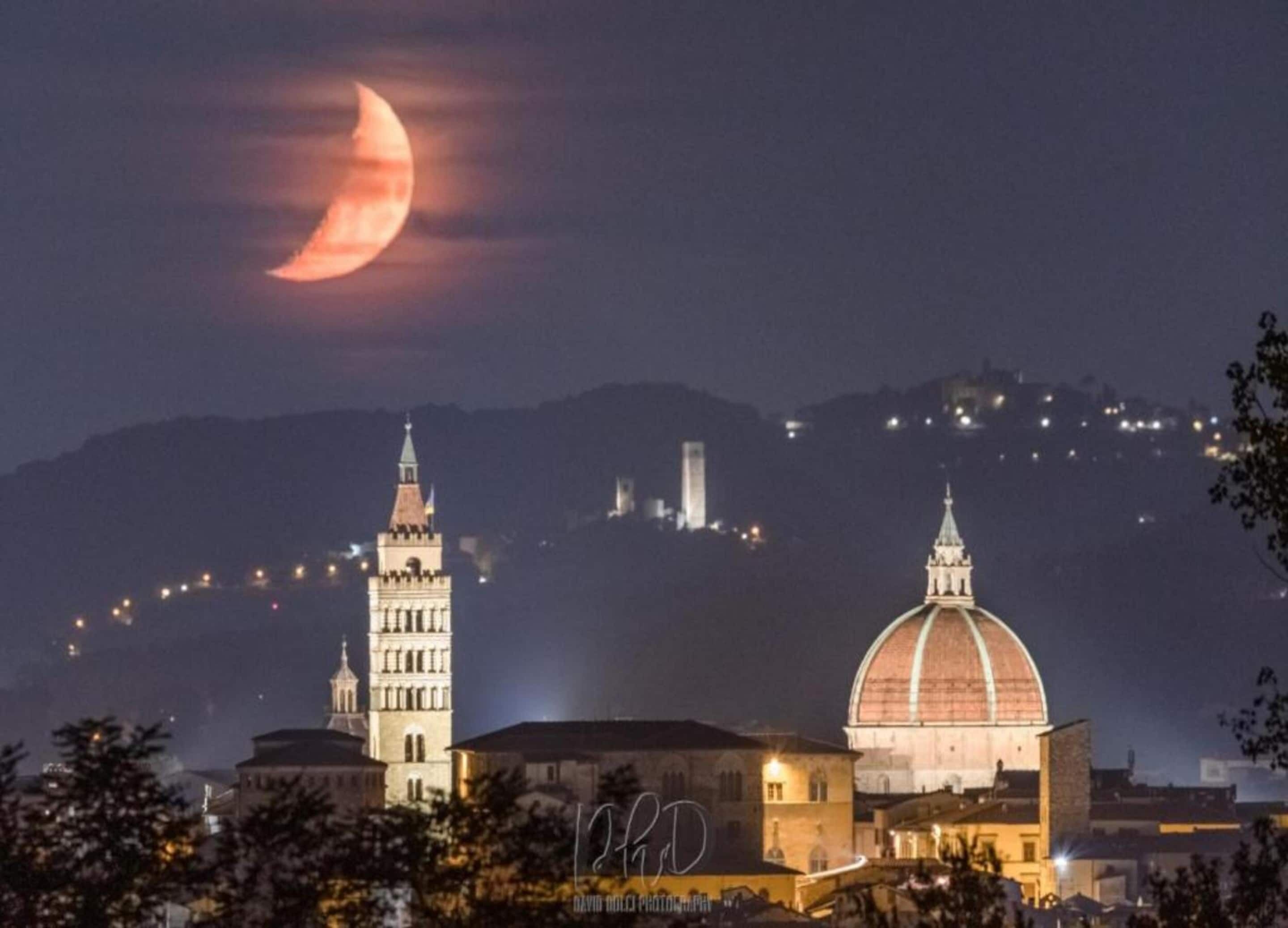 Vista nocturna de un paisaje urbano con una torre destacada y un edificio con forma de cúpula, bajo una luna creciente.