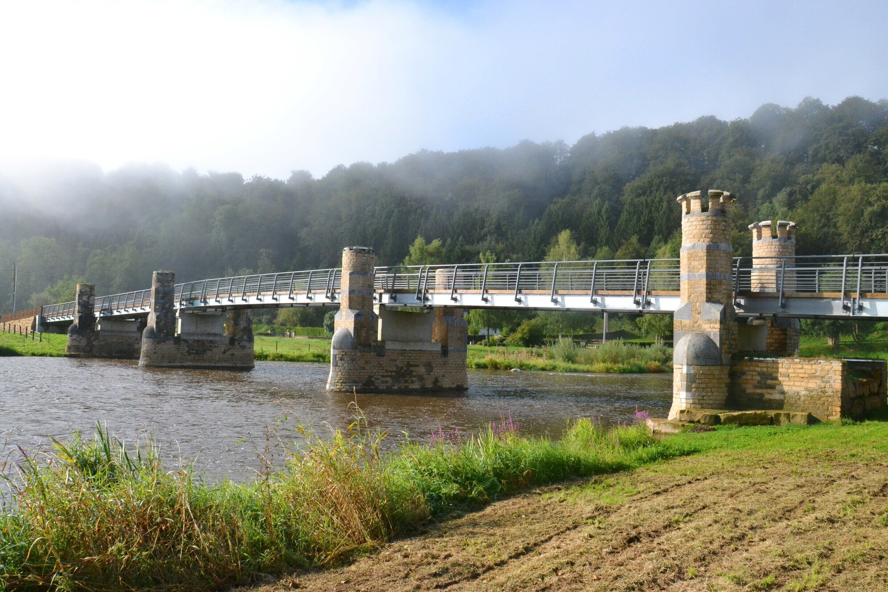 A stone bridge with a metal railing over a river, surrounded by greenery and a forested hill in the background.