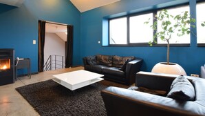 A modern living room with a fireplace, a black leather sofa, a white coffee table, and a large window with a view of the outdoors. - Luxury Cottage in Vaux-sur-Sûre with Hot Tub (Vaux-sur-Sure)
