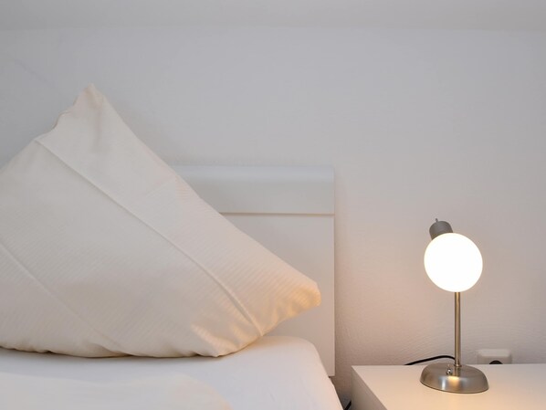 A bed with white bedding and pillows, a bedside table with a lamp, and a white wall. - Wohnung in Willingen Nahe Ettelsberg-bahn (Willingen)