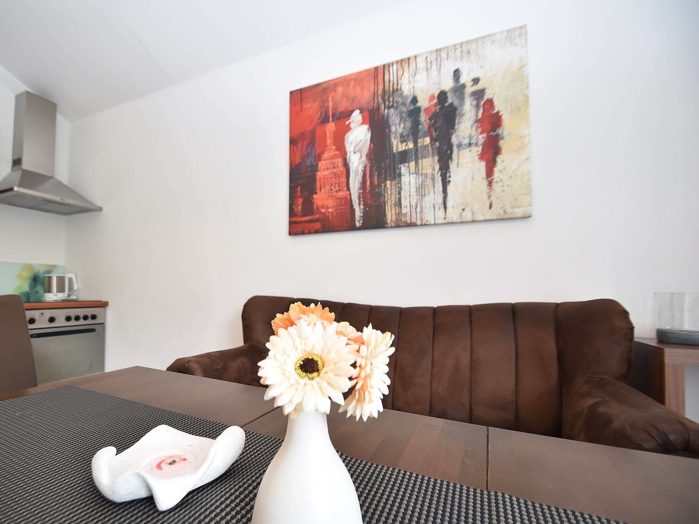A living room with a brown sofa, a white vase with flowers, and a painting on the wall.