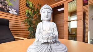 Una statua raffigura una figura seduta che tiene una ciotola, posizionata su un tavolo di legno in una stanza con pannelli in legno e una pianta sullo sfondo.
