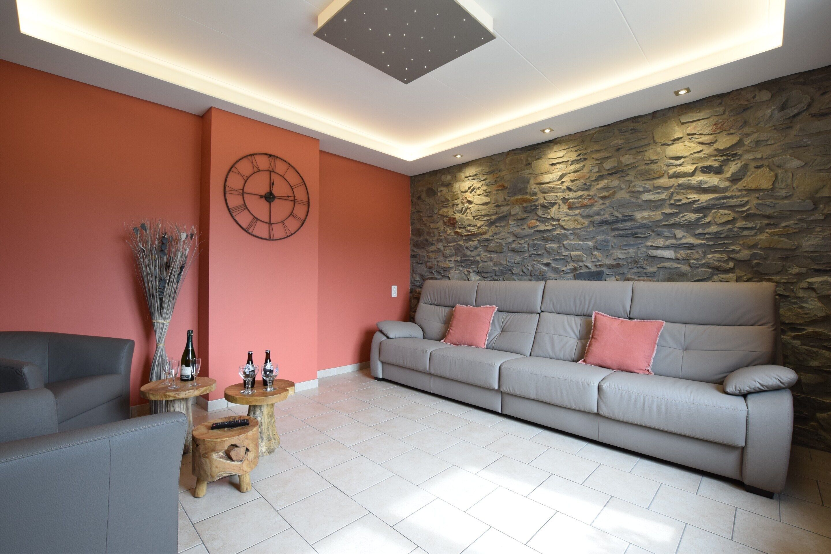 Ein modernes Wohnzimmer mit einer Wand aus Stein, einem grauen Sofa und einem runden Couchtisch mit Weinflaschen.