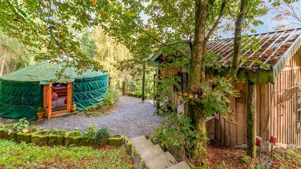 Uma casa rústica de madeira com telhado de palha, um caminho de pedra que leva a uma tenda verde, e uma cerca de madeira.