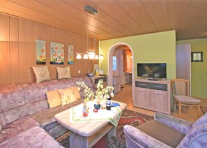A living room with a sofa, coffee table, TV, and kitchen area. - Ferienhaus in Vogtland (Falkenstein/Vogtl.)