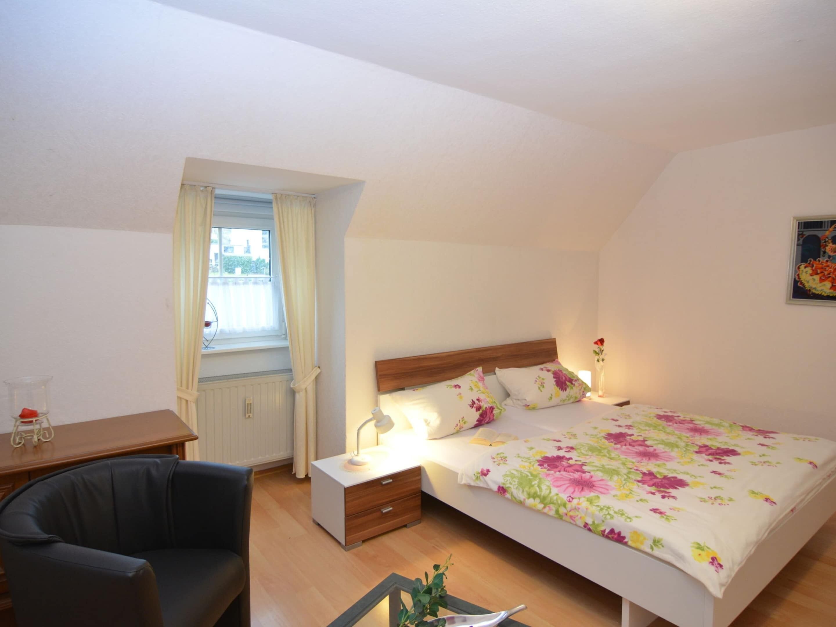Une chambre à coucher comprenant un lit, un bureau, une chaise, une fenêtre avec des rideaux et une table de chevet avec une lampe.
