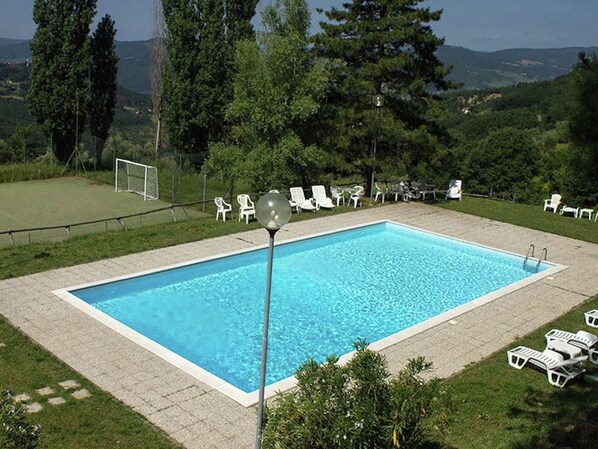 A rectangular swimming pool with a clear blue water surface, surrounded by a paved deck. There are several white lounge chairs arranged around the pool, and a soccer goal is visible in the background. - Belvilla by OYO Santa Cecilia (Rufina)