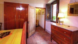 A bedroom with a wooden wardrobe, a bed with a striped bedspread, a dresser with drawers, a mirror, and a window with a view of greenery. - Belvilla by OYO Santa Cecilia (Rufina)
