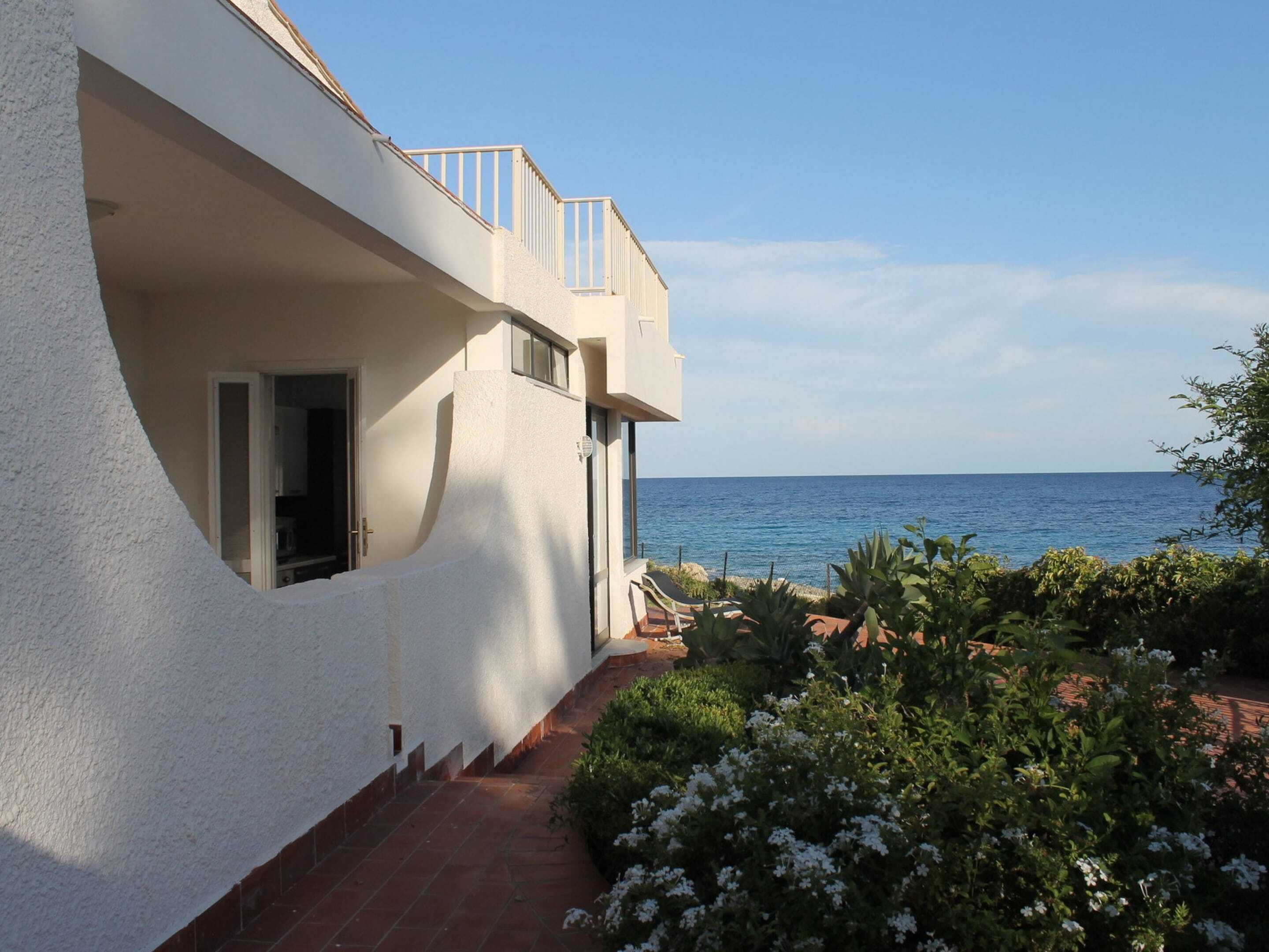 A white building with a balcony overlooking the sea, a paved path, and flowering plants.