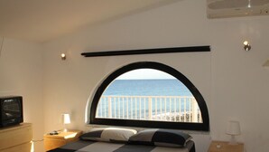 A bedroom with a checkered bedspread, a television on a wooden cabinet, a bedside lamp, and a view of the sea through a large arched window. - Belvilla by OYO Villa Eirene (Syracuse)