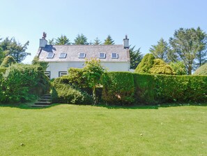 A house with a white exterior, grey roof, and multiple chimneys. There is a well-maintained lawn in front, and a hedge to the right.