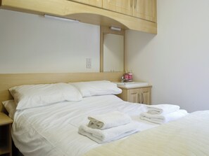 Un lit bien fait, avec du linge de lit et des serviettes blanches, une tête de lit en bois, une petite table de chevet avec un miroir et des articles de toilette, et un mur de couleur claire.