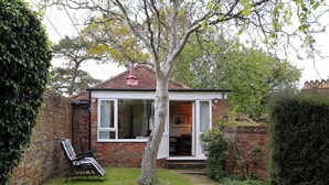Una pequeña casa de ladrillo con una ventana blanca y una puerta, un árbol al frente, y una silla con cojín a rayas.
