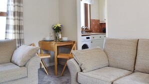 A cozy living room with a sofa, a wooden dining table with a vase of flowers, and a kitchen visible through an open door.