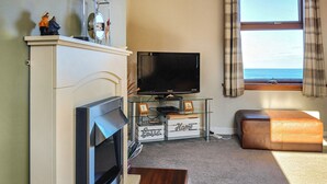A living room with a fireplace, a television, a glass coffee table, and a view of the sea through a window.